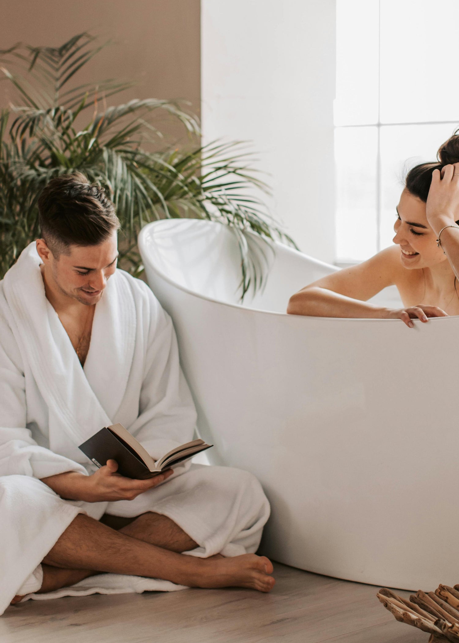 Man reading a book next to a woman in a bathtub in a modern bathroom.